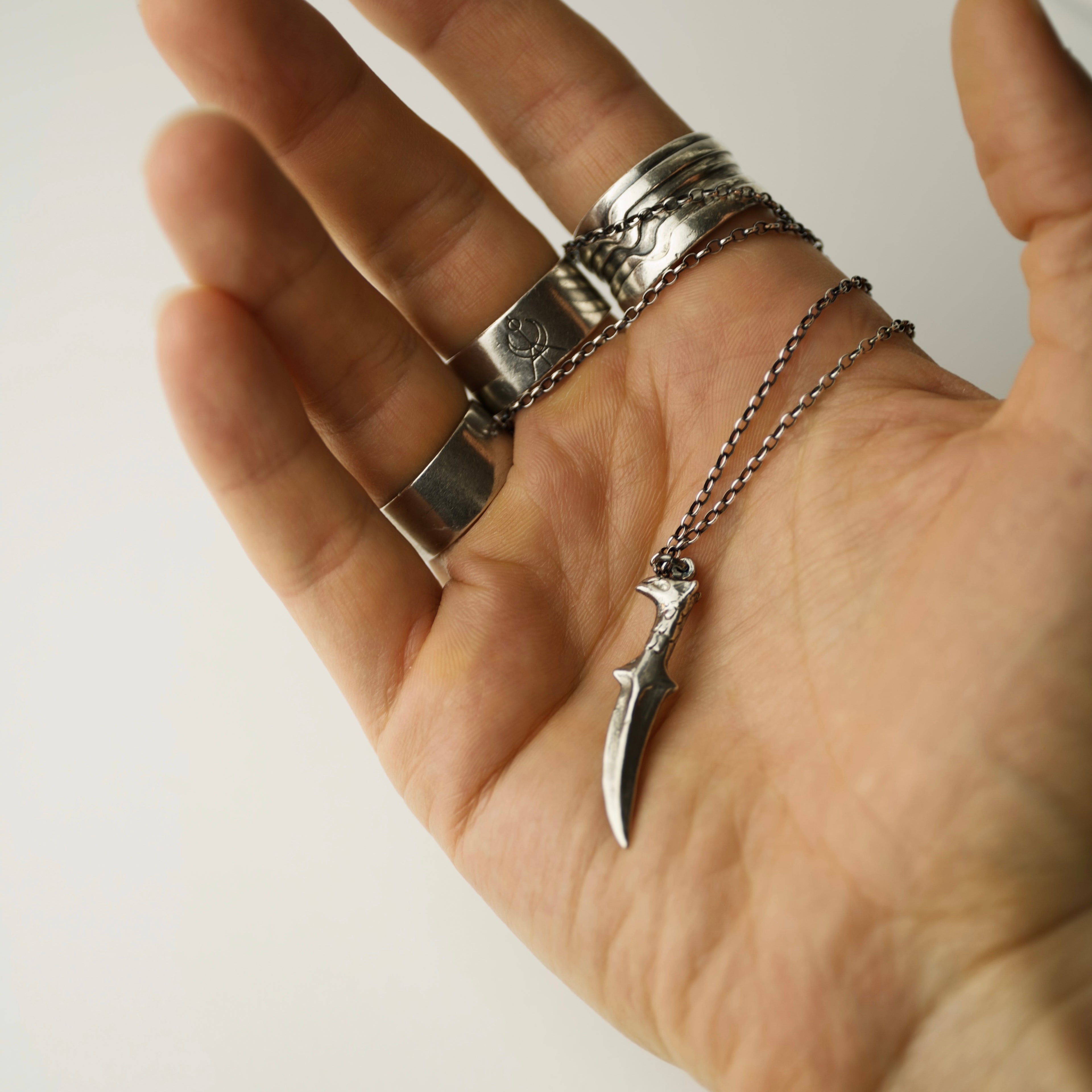 Hand wearing multiple silver rings by Macabre Gadgets holding a Griffin Sabre pendant with silver chain on a plain background