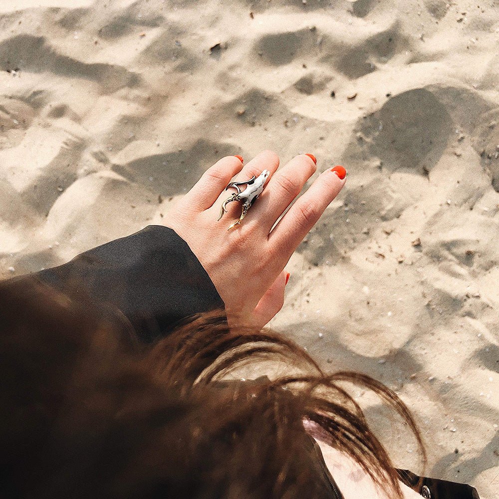 Doe Skull ring on a woman's hand with crimson nails, we can see few locks of brown hair and sand beach, cinematic yet lifestyle image