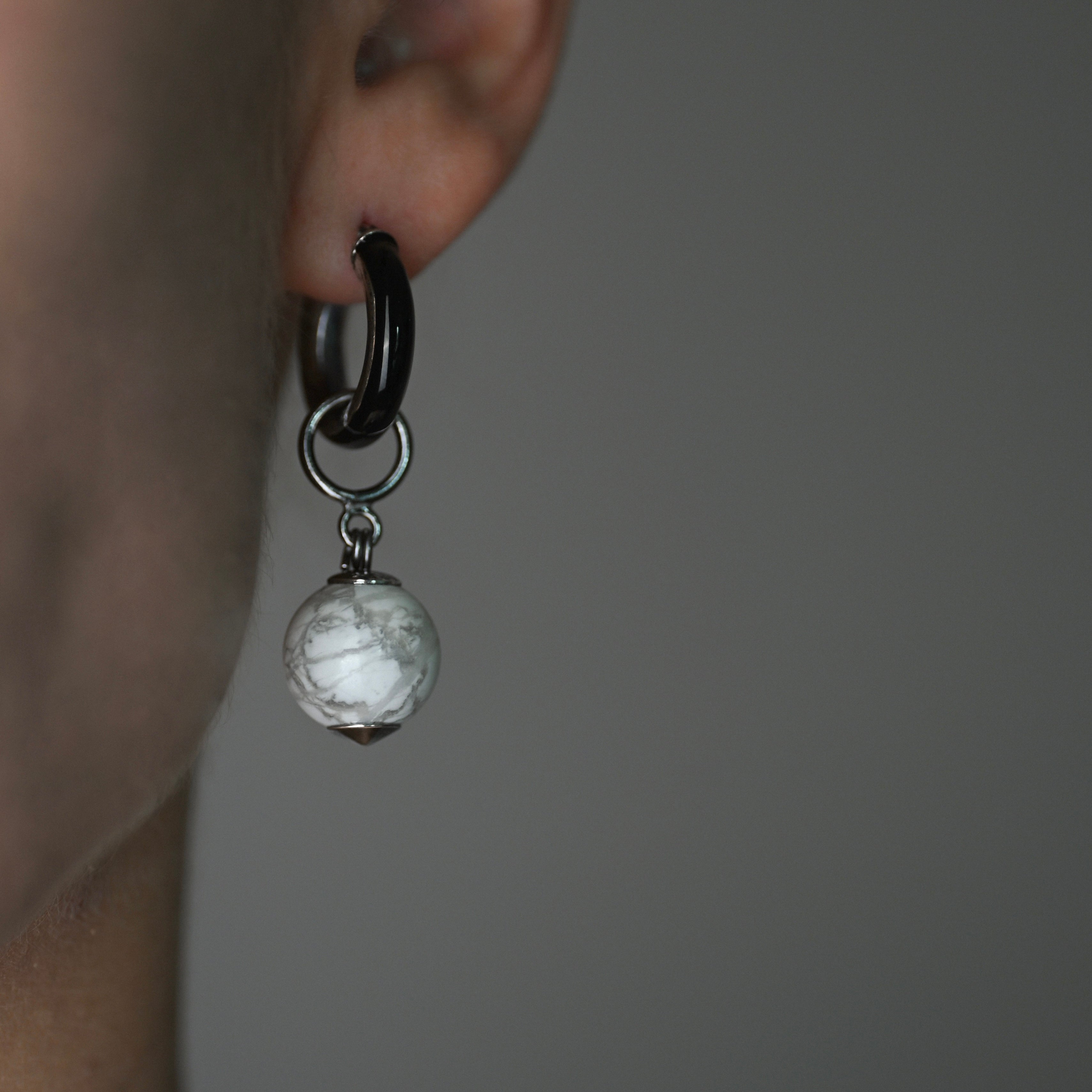 Close-up of an earring with a dark band and a round, light-colored gemstone on a neutral background.