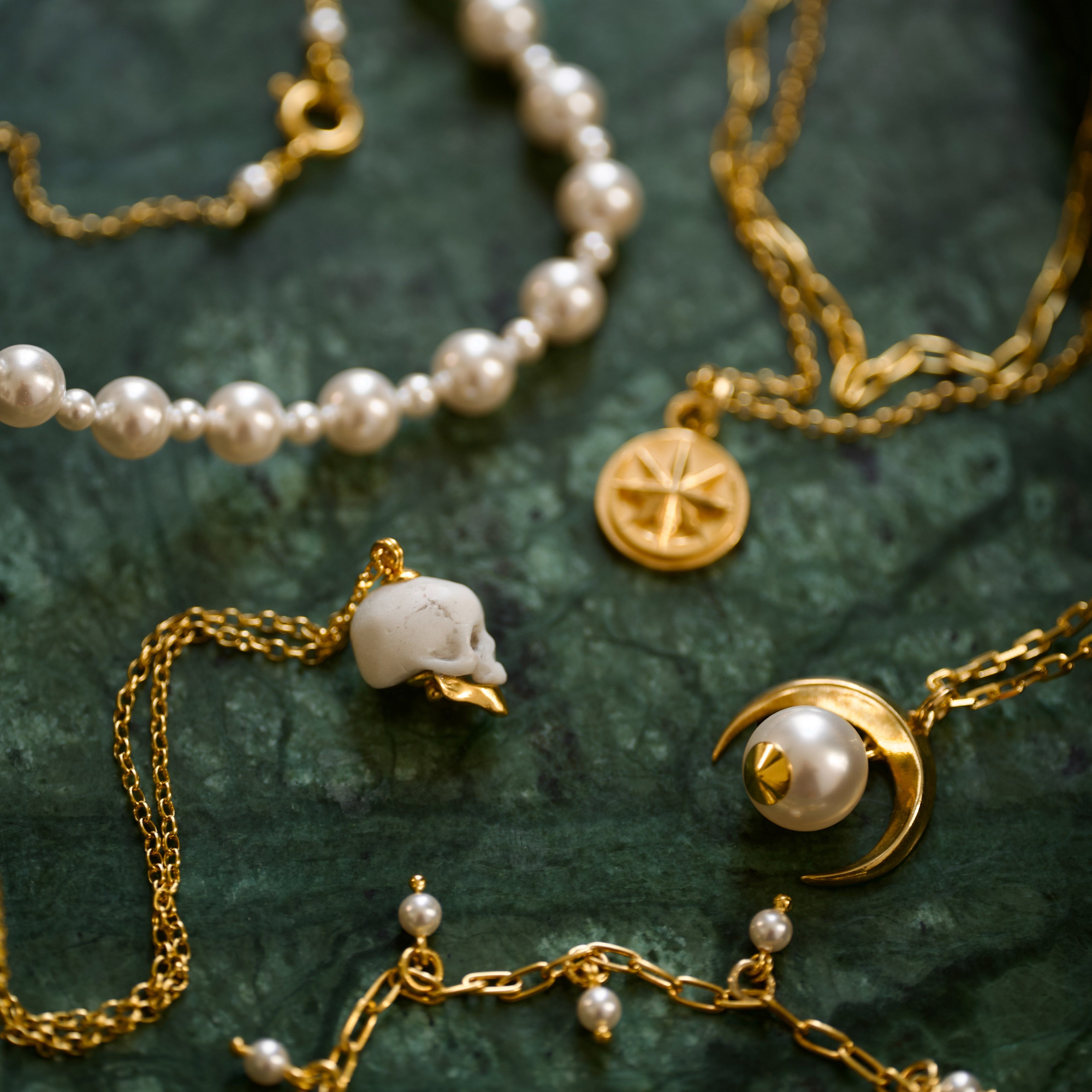 Ancient Skull pendant, Aster pendant and other gold and pearl necklaces by Macabre Gadgets on a marble, dark green surface