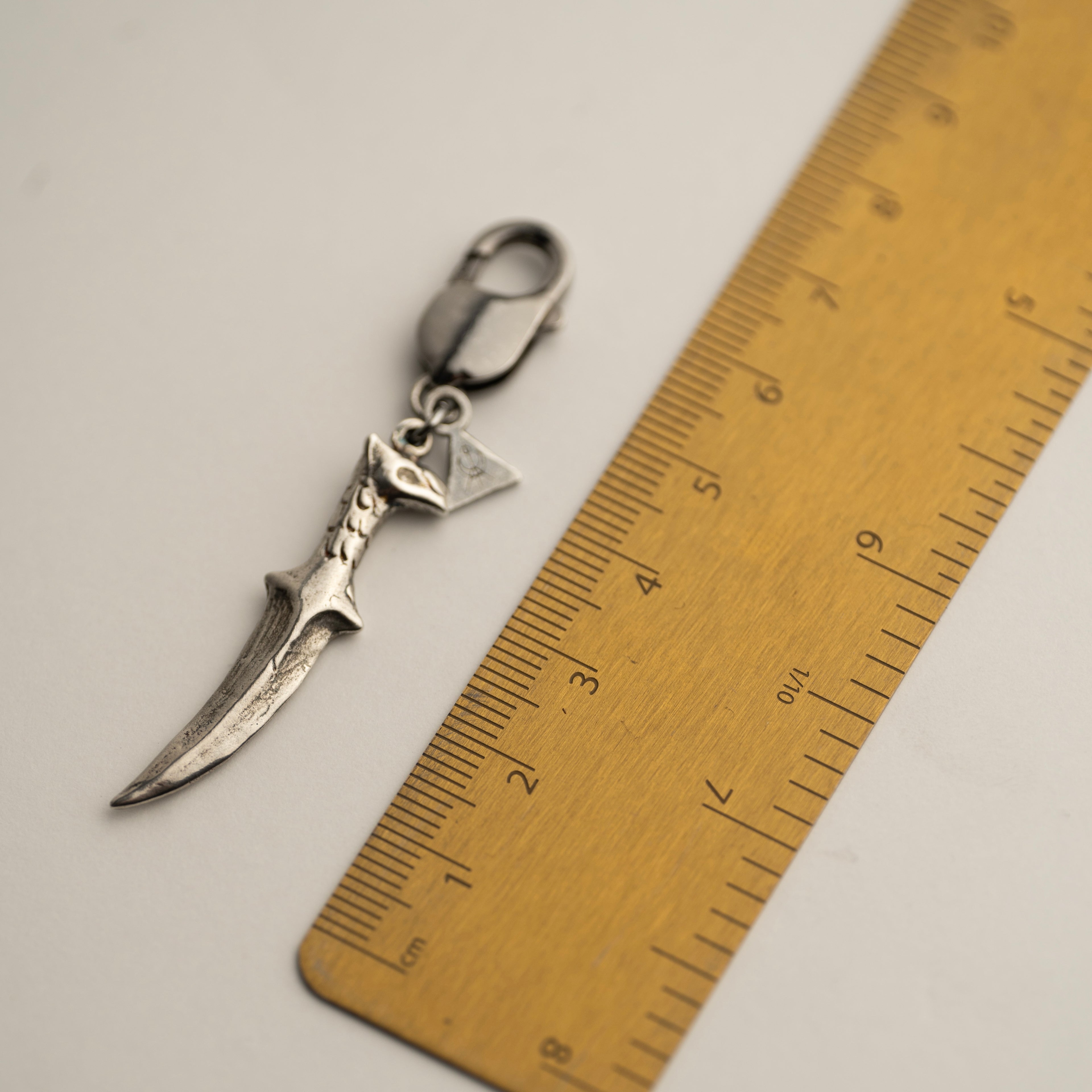 Silver dagger-shaped key-chain next to a ruler on a light gray background to show overall size of the piece