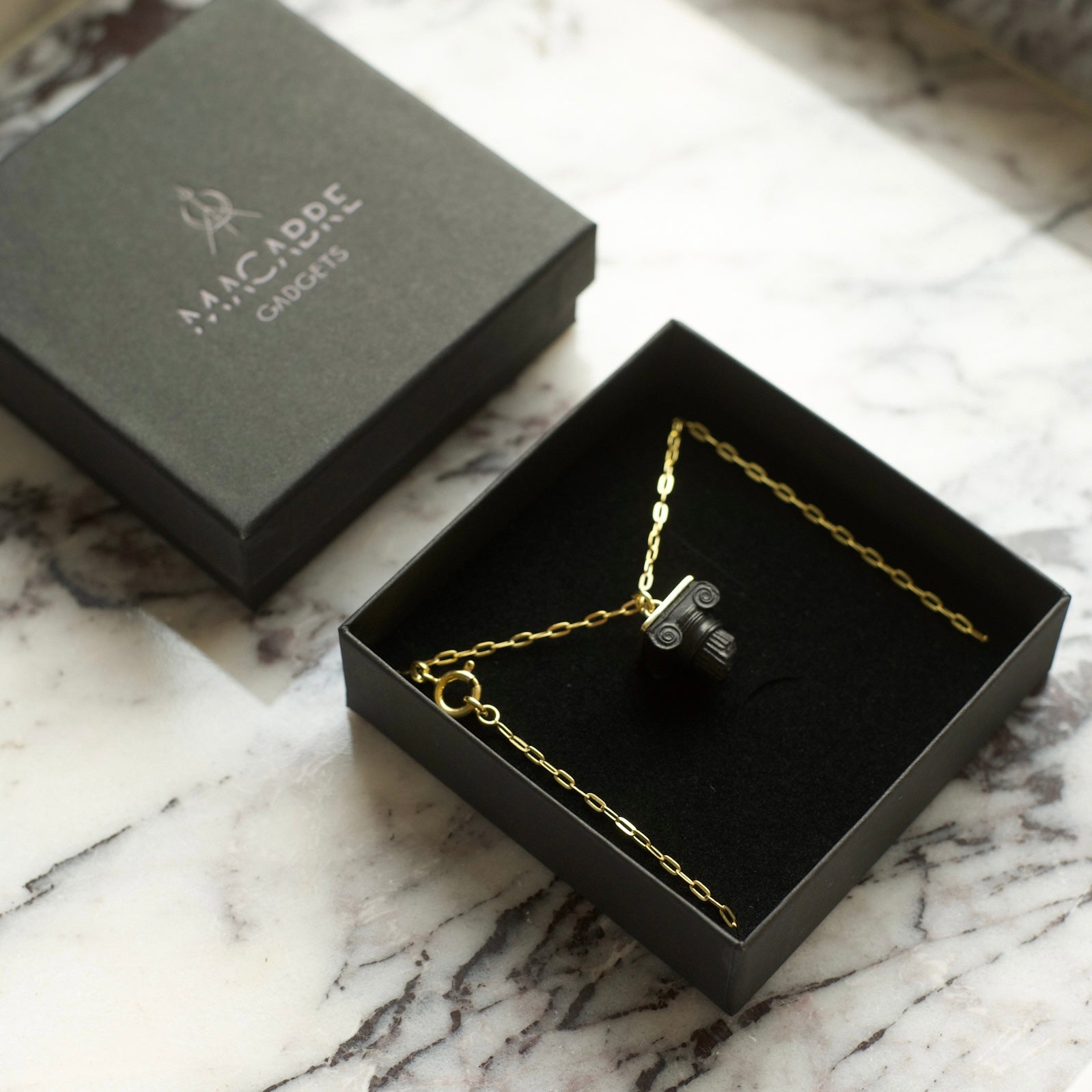 Black pendant in a form of a miniature Ionic column capital with 24k gold plated anchor chain in a luxurious black box on a marble surface - minimalistic lifestyle shot