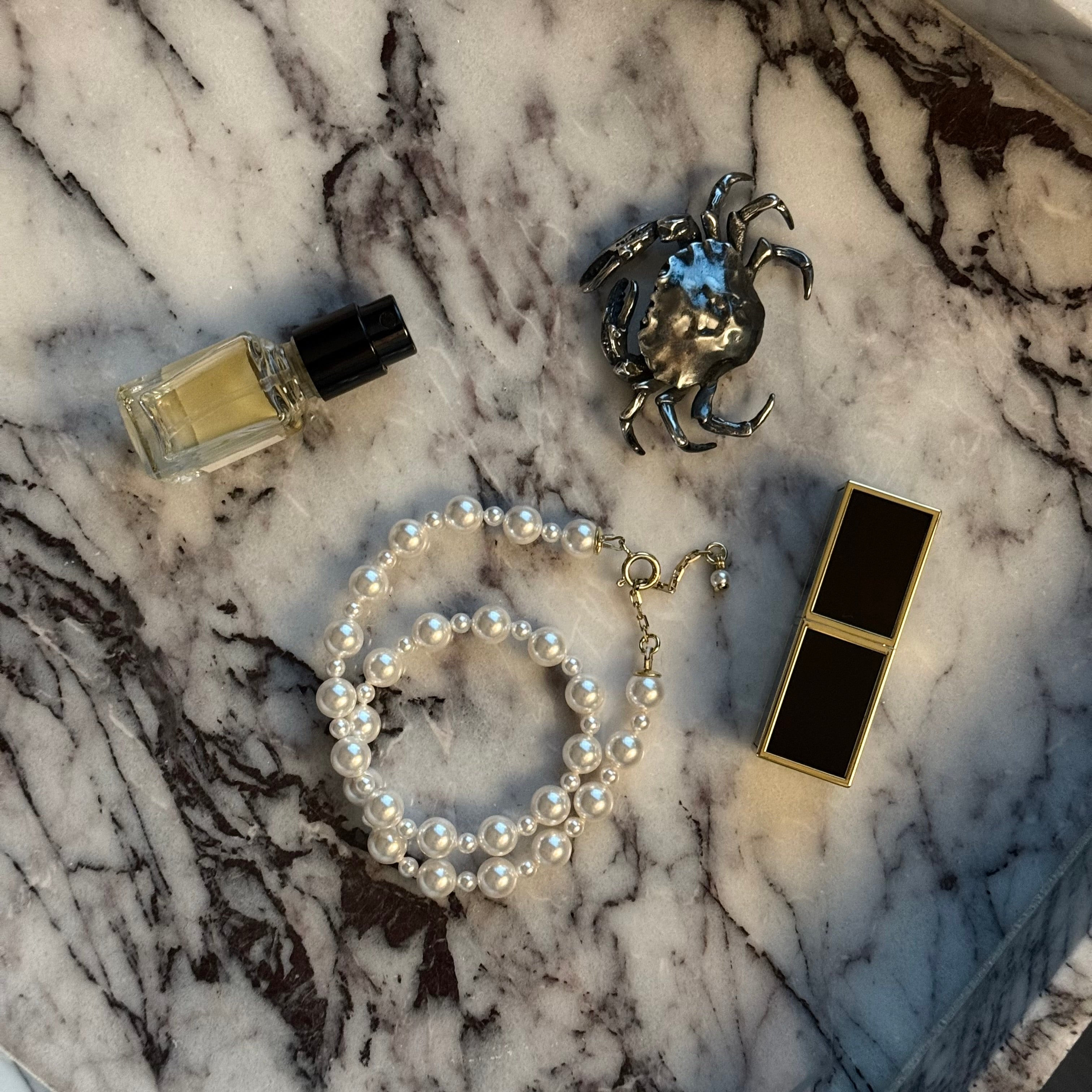 Macabre Gadgets Pearl necklace, silver crab-shaped brooch, and makeup compact lipstick on a marble surface