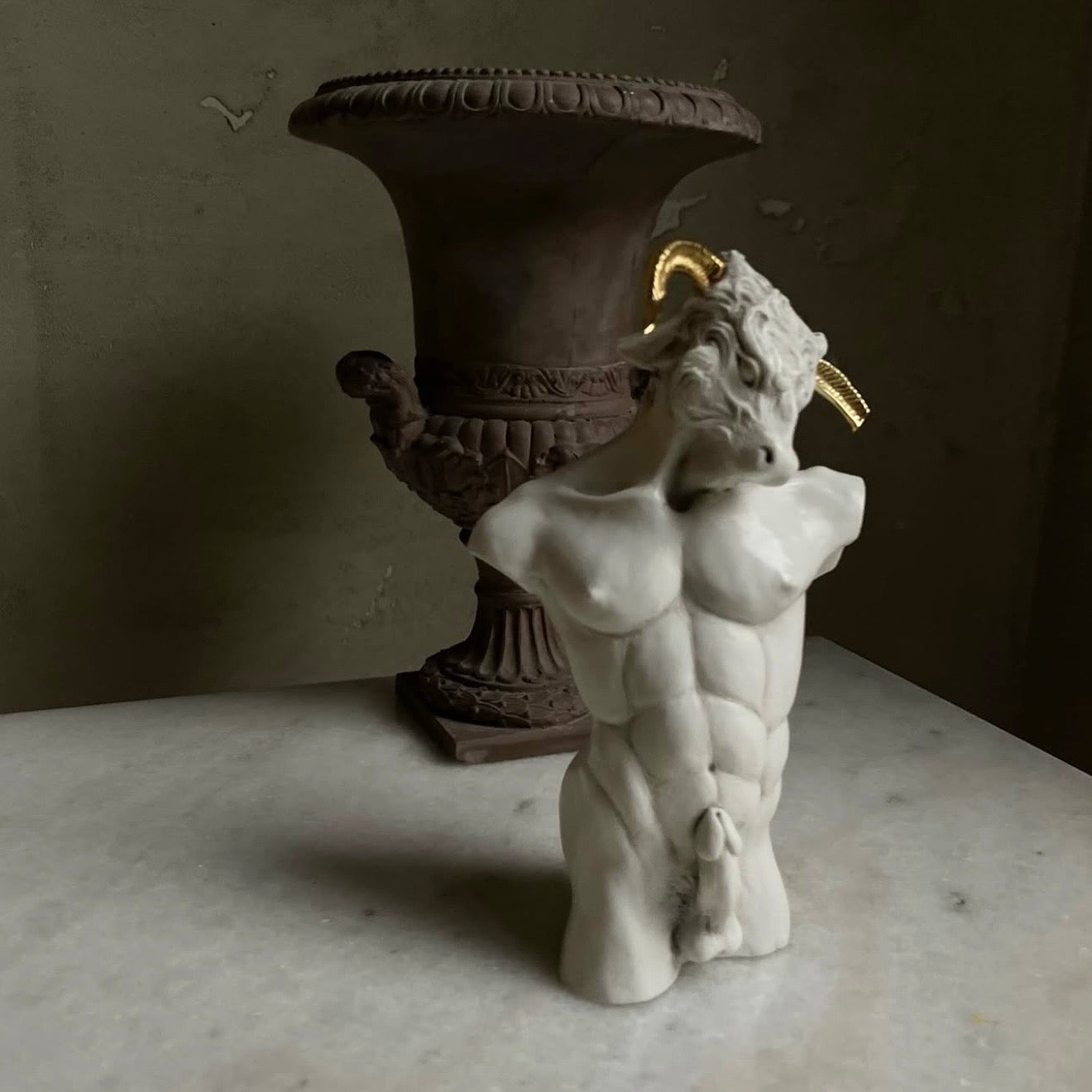Marble sculpture of a figure of Minotaur  with a gold horns in front of an ornate brown Classical vase.