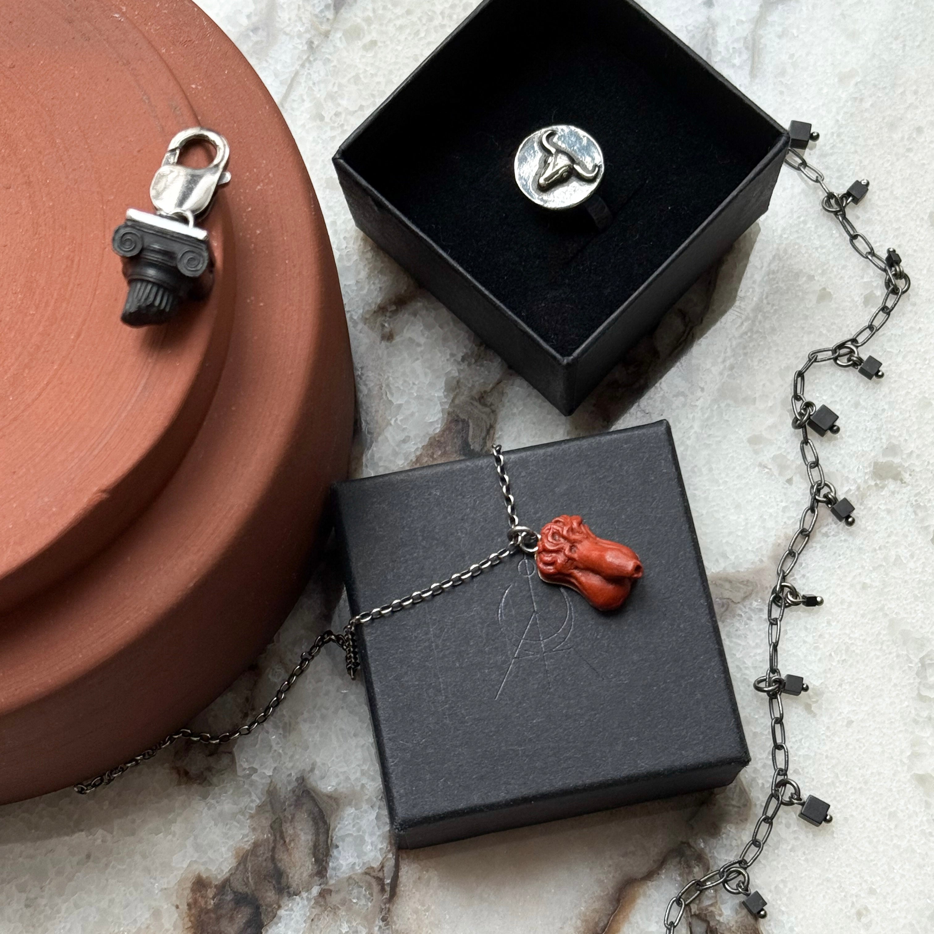 Lifestyle picture of Ionic Charm in black color, Bull Ancient Coin ring in a black box, Phallus pendant in terracota and Mosaic necklace in black and silver, all on marble and terracota background