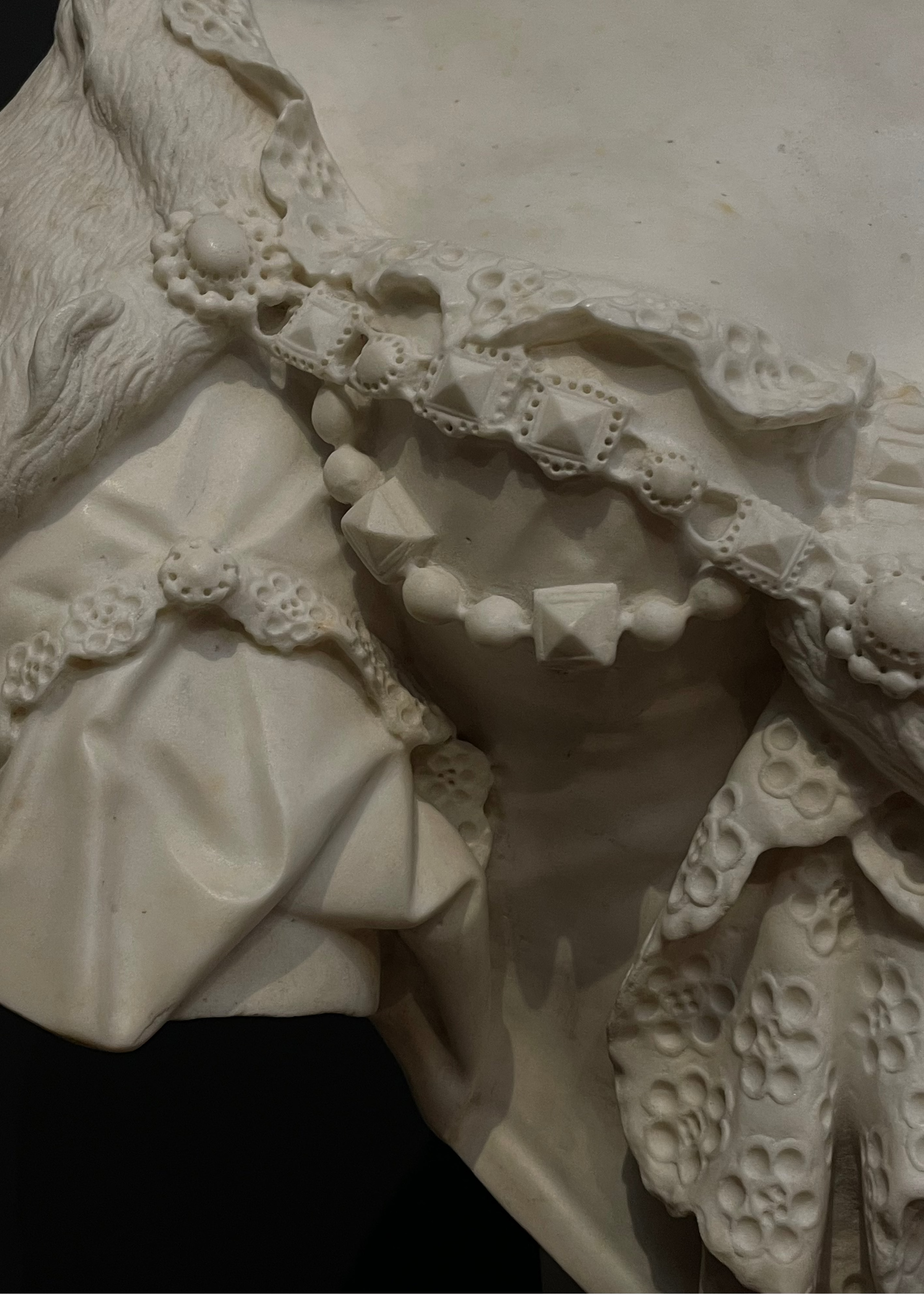 Close-up detail of an off-white carved decorative garment or sculpture with layered ruffles, lace-like trim, and sculpted floral patterns against a dark background.