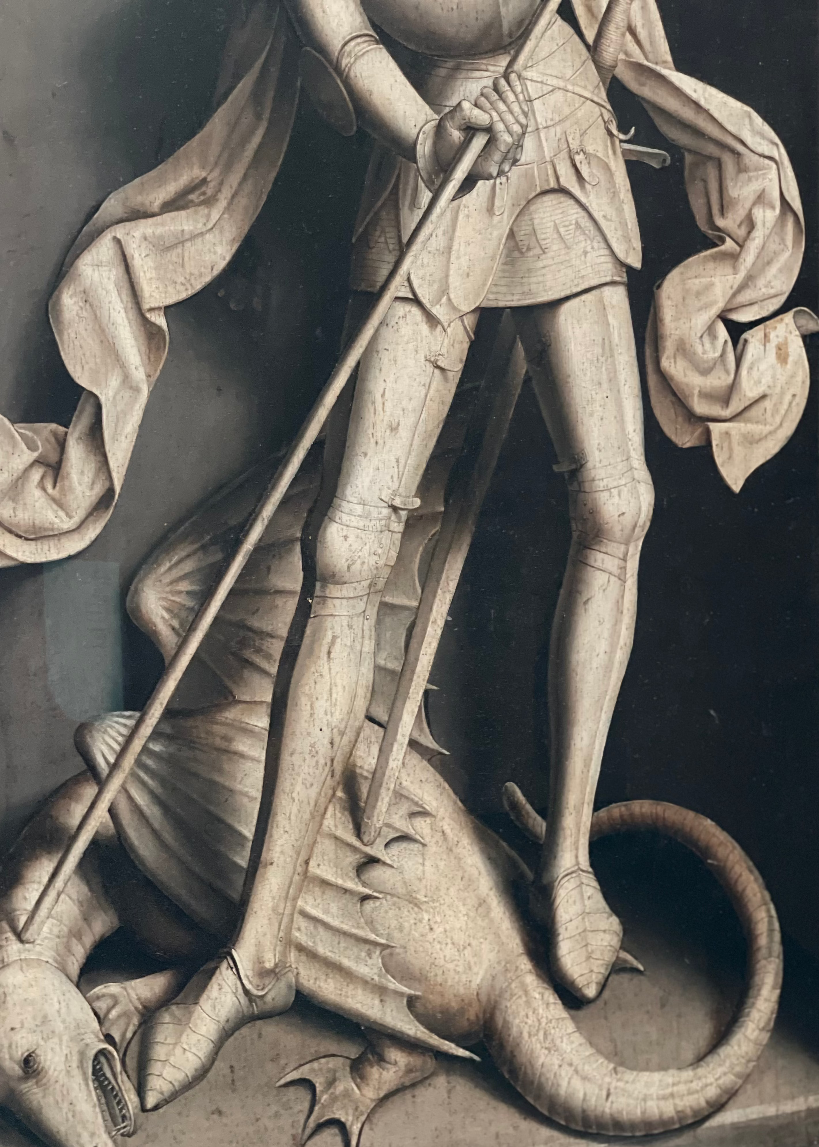 A close-up of a painting of a sculpture depicting a knight in chainmail and a helmet standing on a dragon, holding a spear. The carving shows detailed textures and weathered tones.