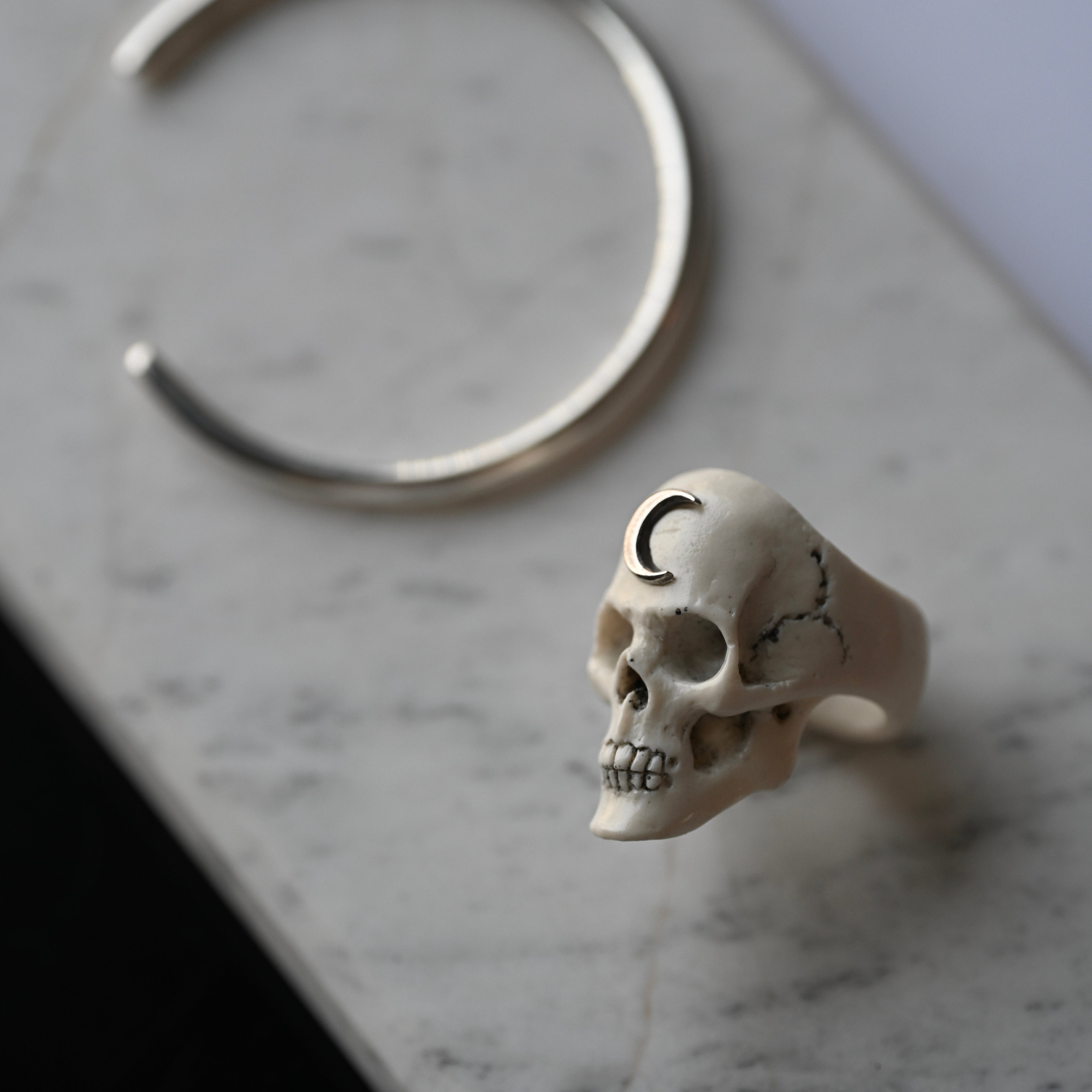 Skull-shaped ring with a crescent moon detail and sterling silver cuff bracelet on a marble surface 