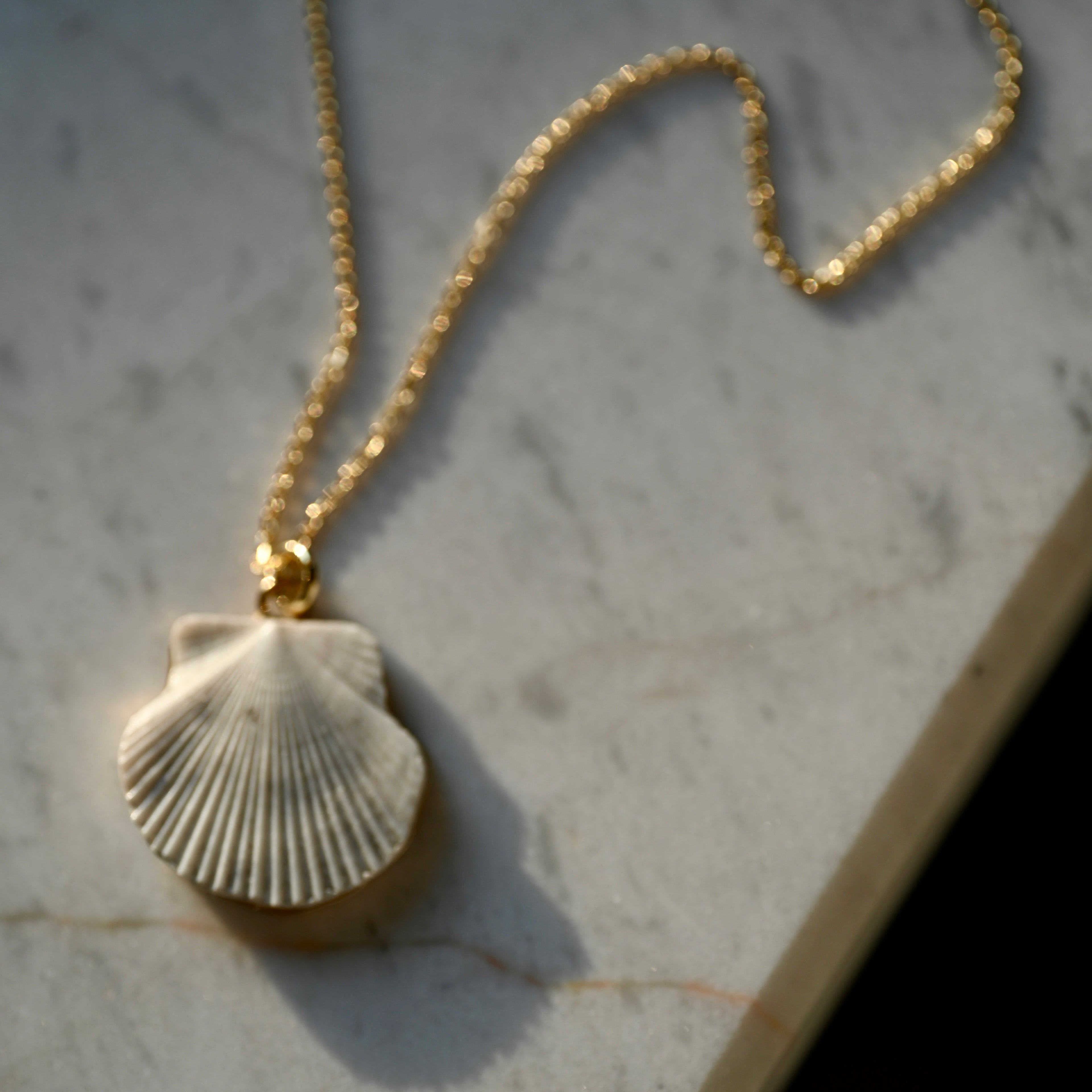 Gold necklace with a shell shaped pendant handcrafted by Macabre Gadgets on a marble surface under contrast light.