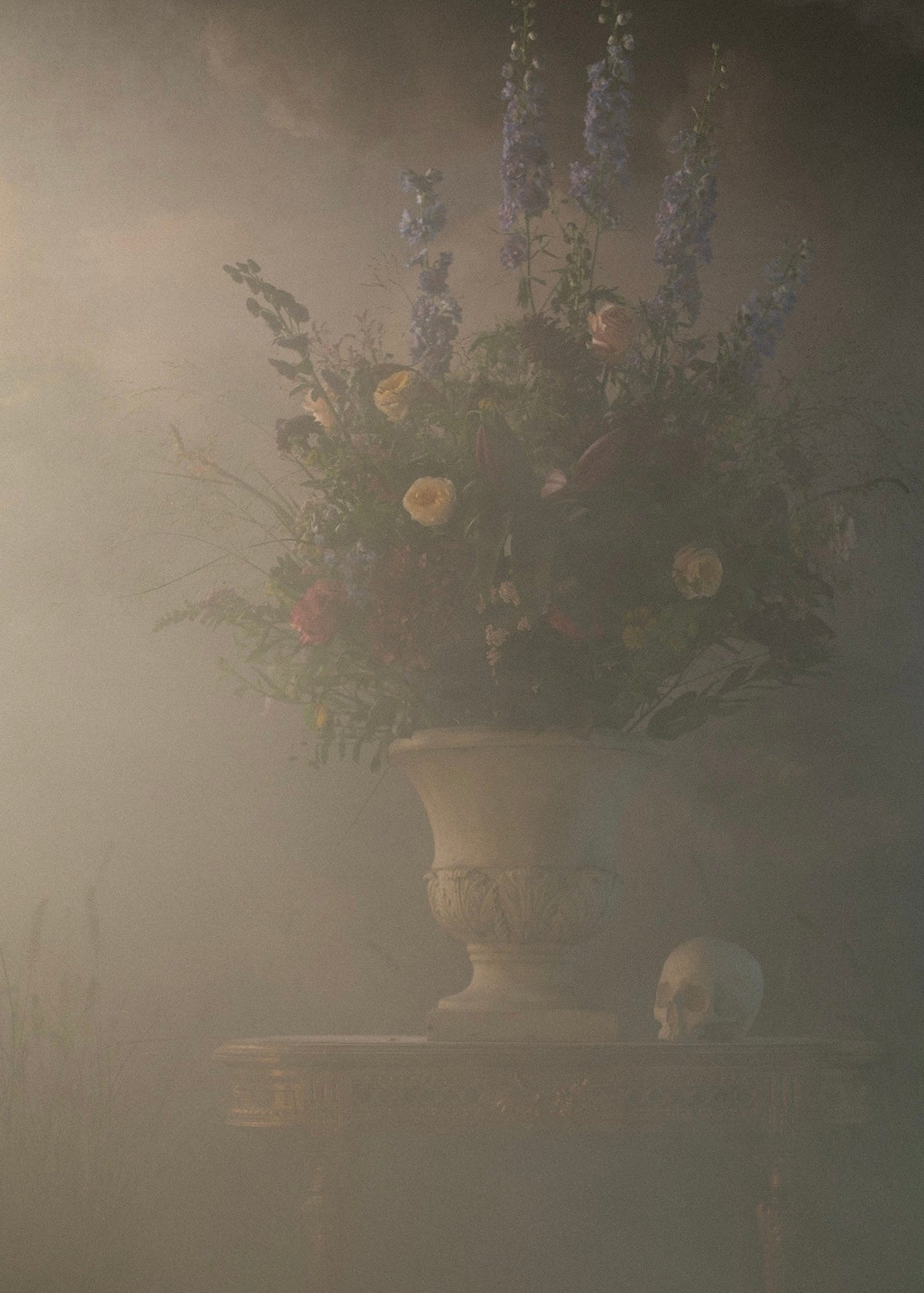 Storm editorial — a moody still life by Macabre Gadgets: a large urn-shaped vase filled with a dense floral arrangement in muted purple, cream, green and red tones, set on a gilded Rococo table beside a white skull vanitas in a foggy atmospheric setting