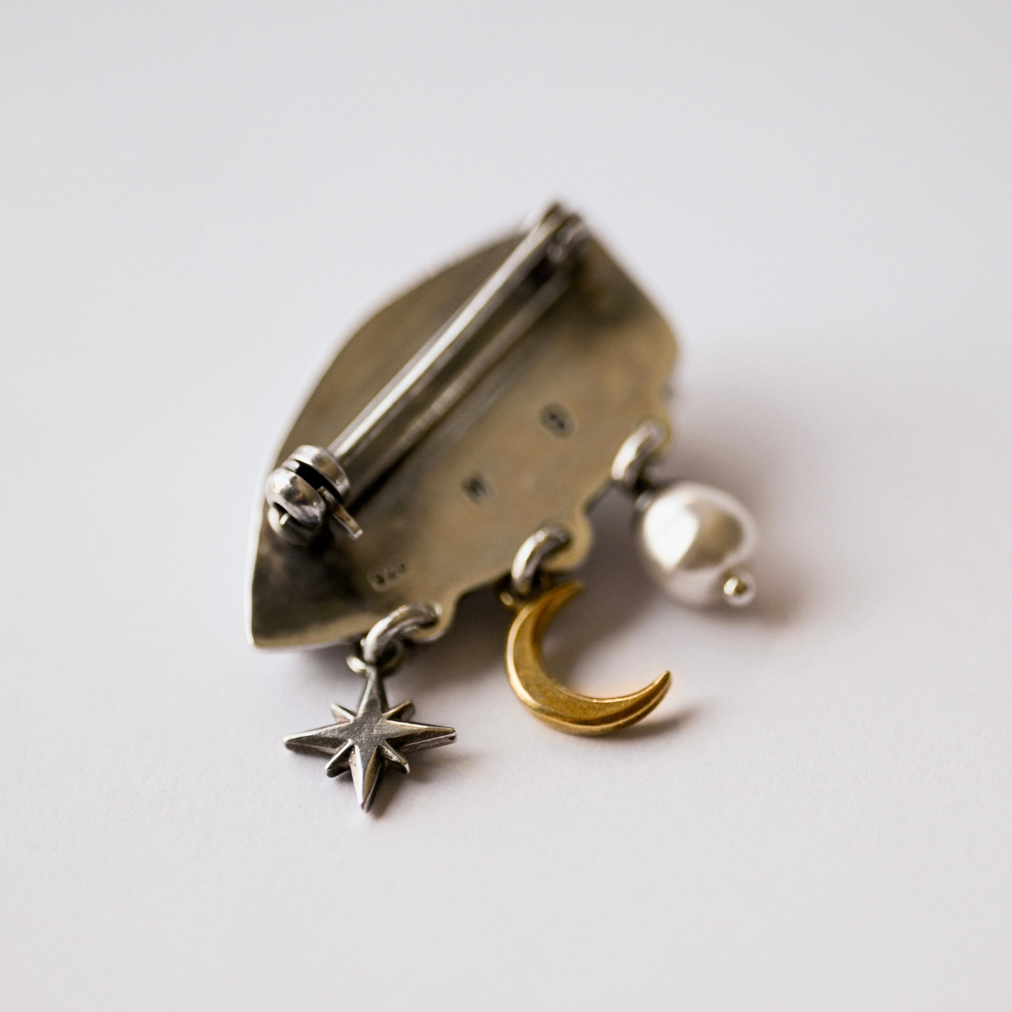 Gaze brooch by Macabre Gadgets - view from the back showing brooch fixture pin and celestial pendants: silver star, golden moon and a pearl on a light background.