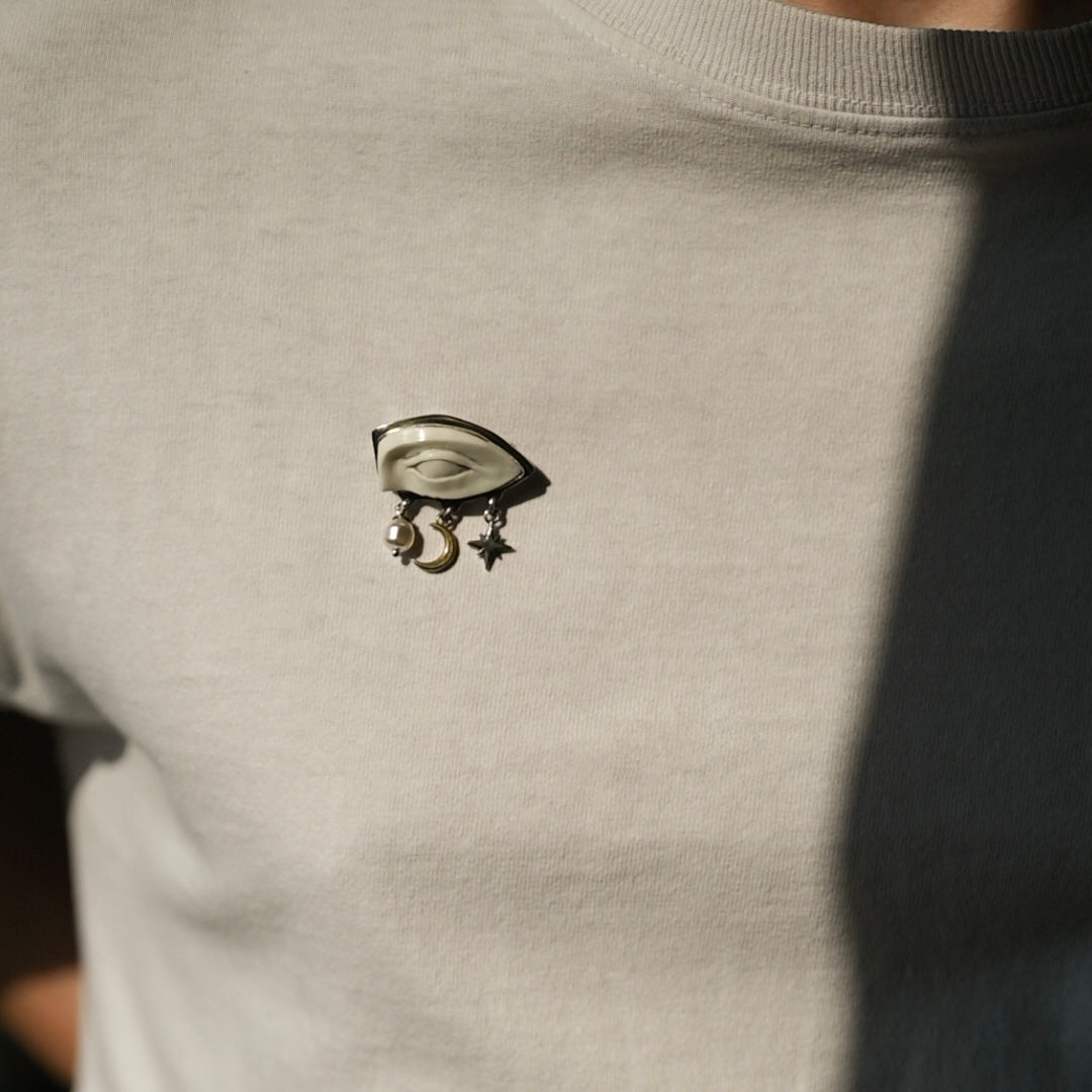Macabre Gadgets Gaze brooch: a white marble eye of a statue with star, moon and pearl dangle charms on a white t-shirt