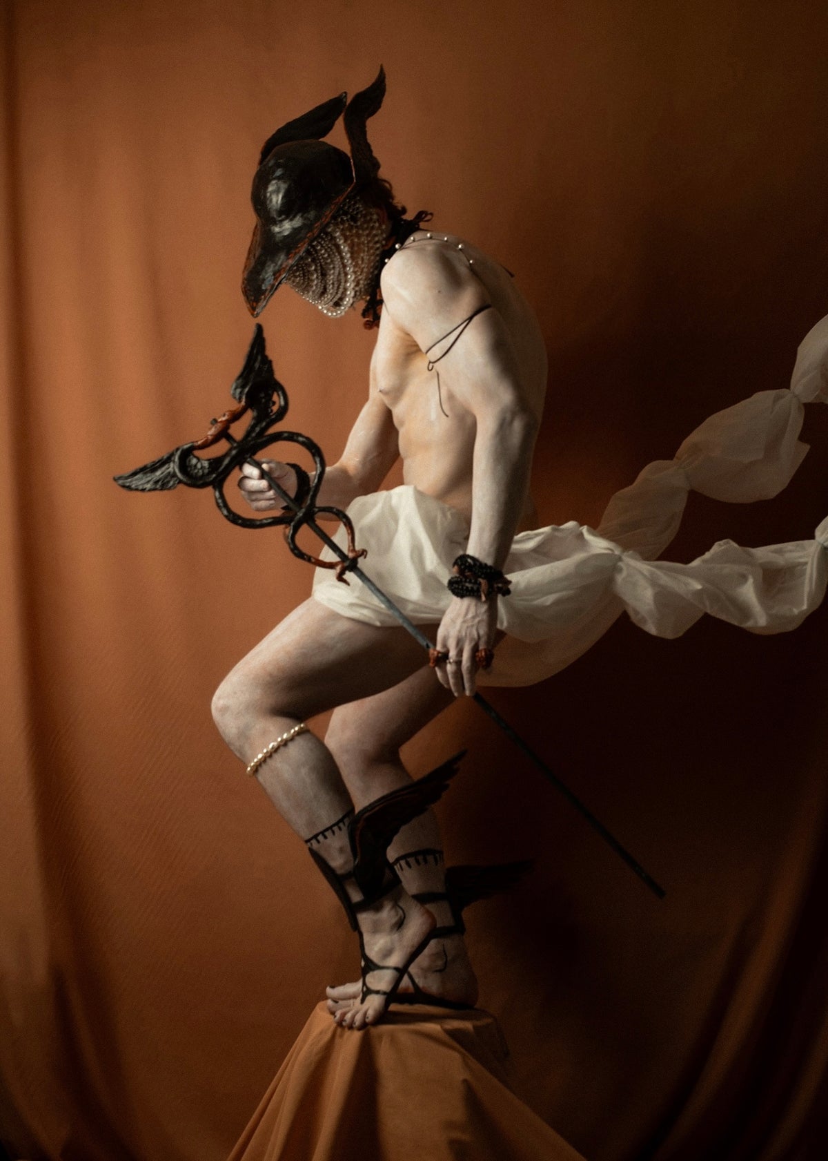 Gods and Monsters campaign — Hermes in cloud-white drapery and caduceus against terracotta, the trickster god between desire and death, THEA by Macabre Gadgets
