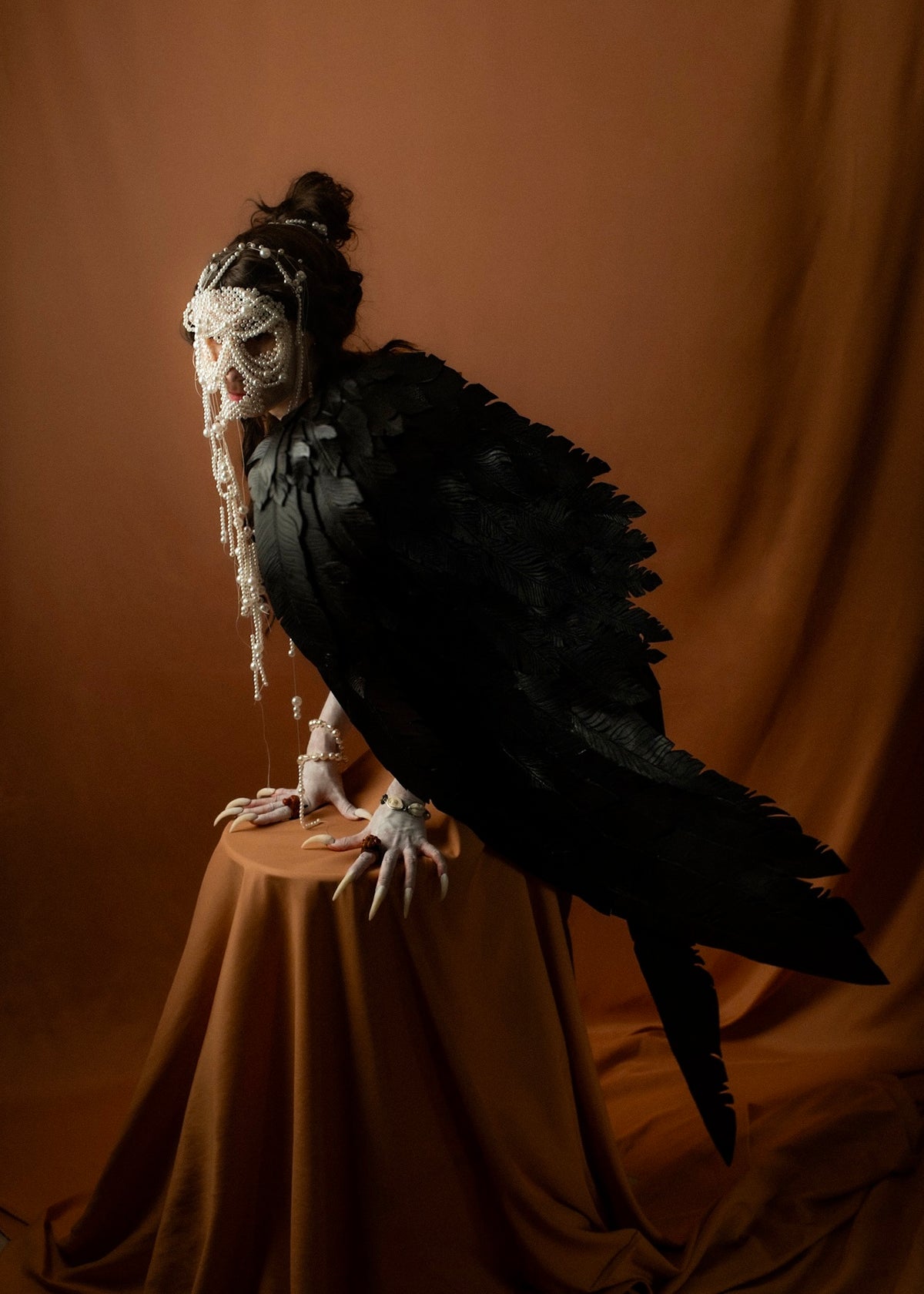Black-feathered Siren seated on a pedestal against a terracotta background, inspired by figures on Ancient Greek pottery, Gods and Monsters campaign | Macabre Gadgets