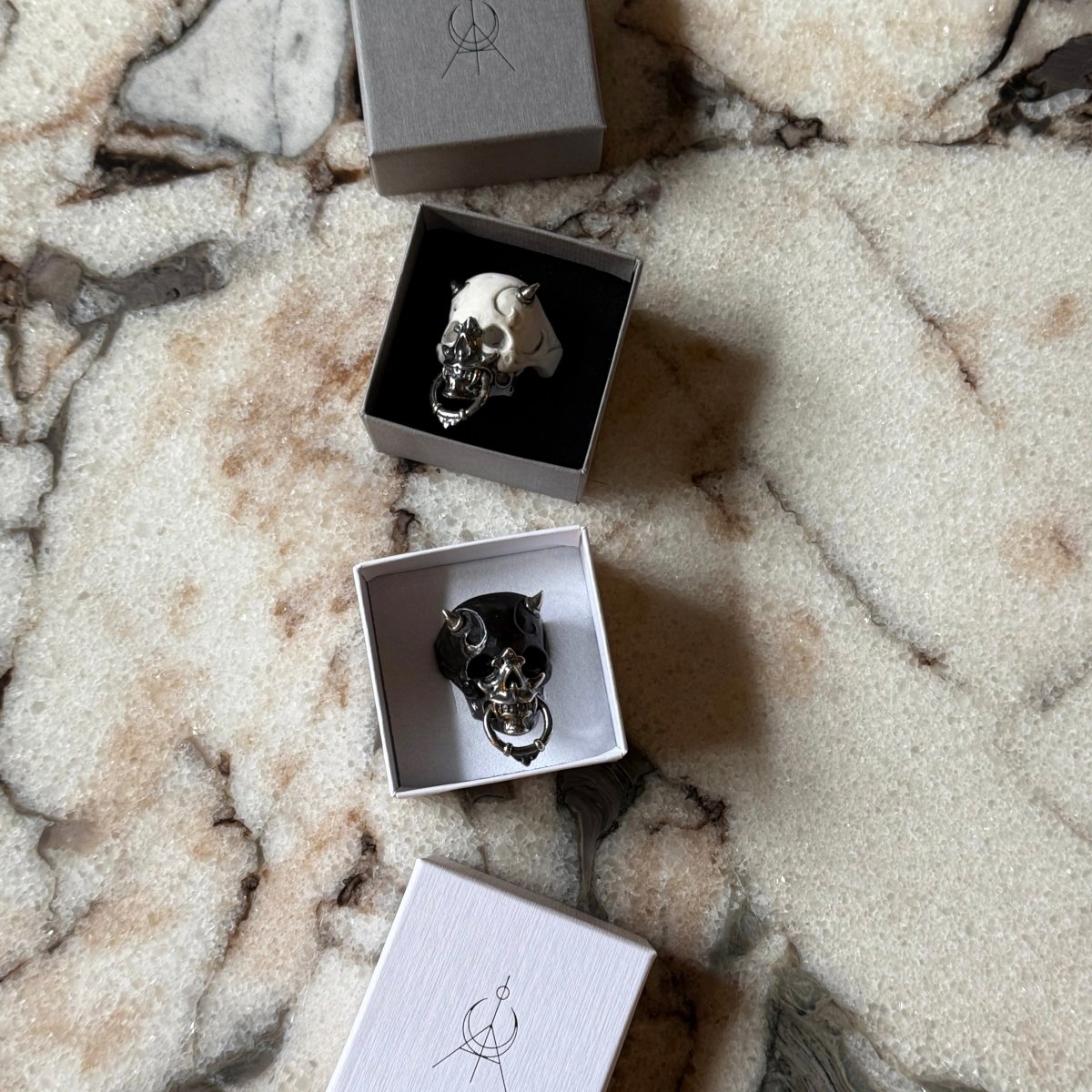 Masked Skull ring — white and black marble variants — in Macabre Gadgets jewelry boxes on a white marble surface