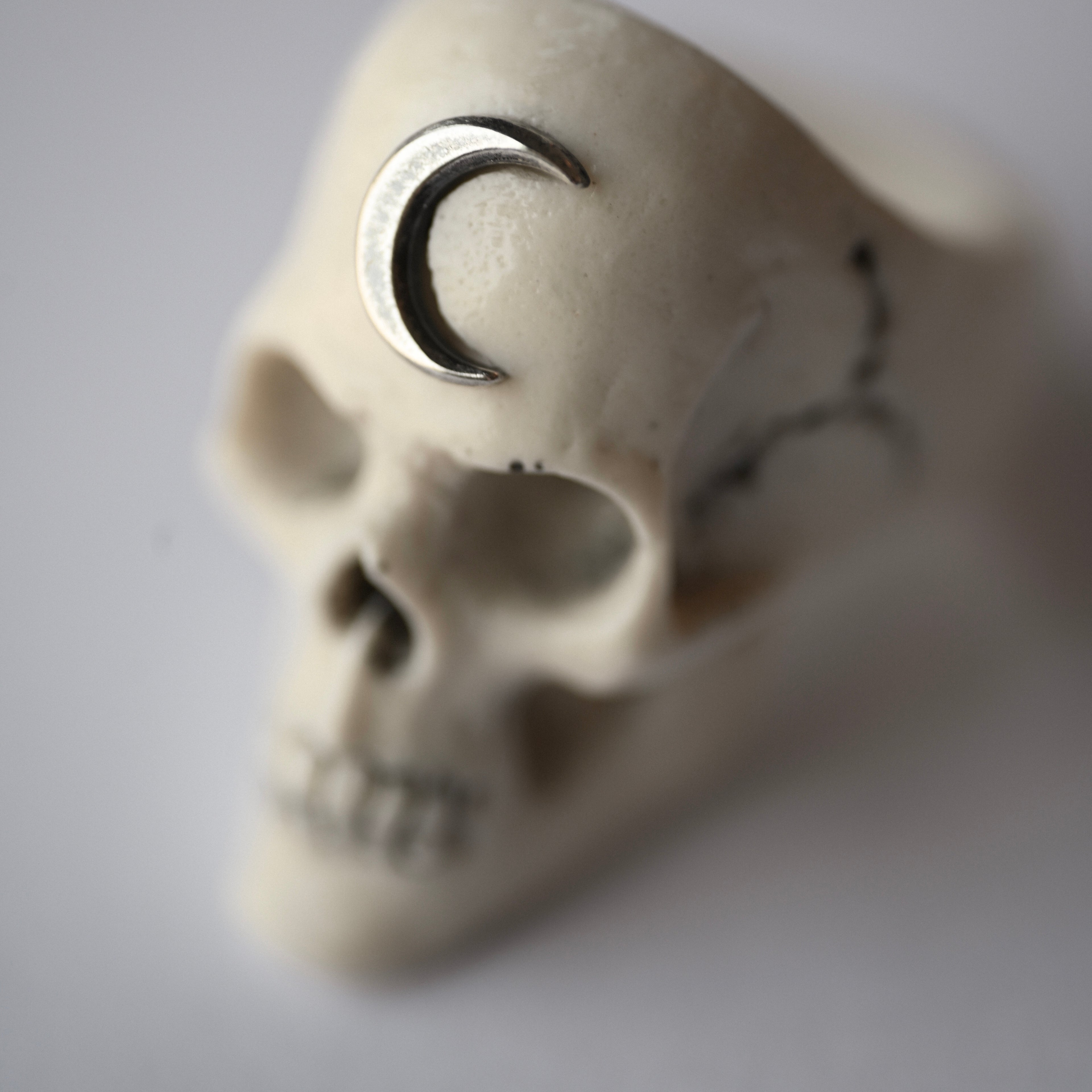 Skull-shaped white marble ring hand crafted by Macabre Gadgets with a silver crescent moon on its forehead on a light gray background