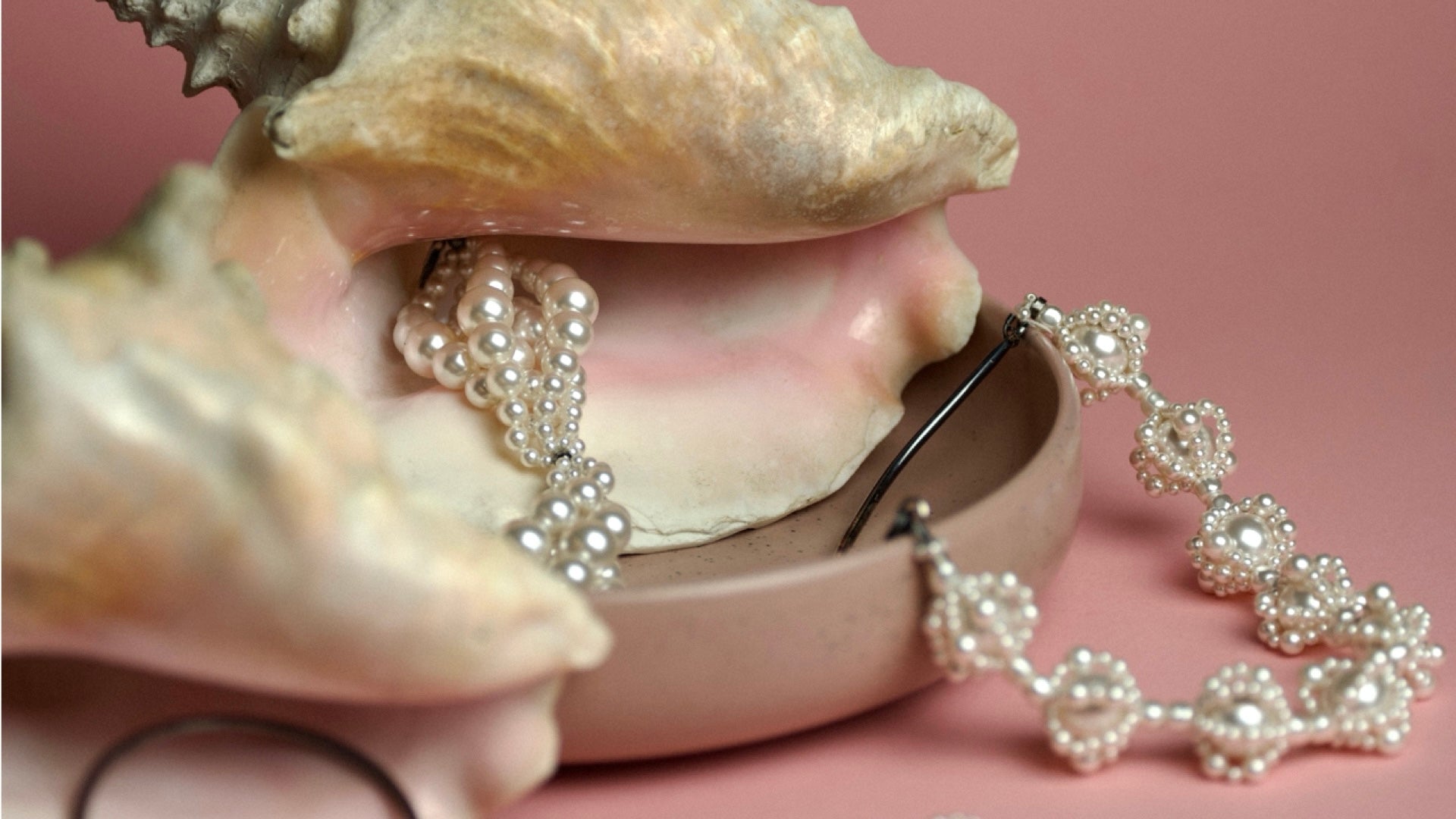 Pearl necklace and bracelet draped over a seashell on a pink background
