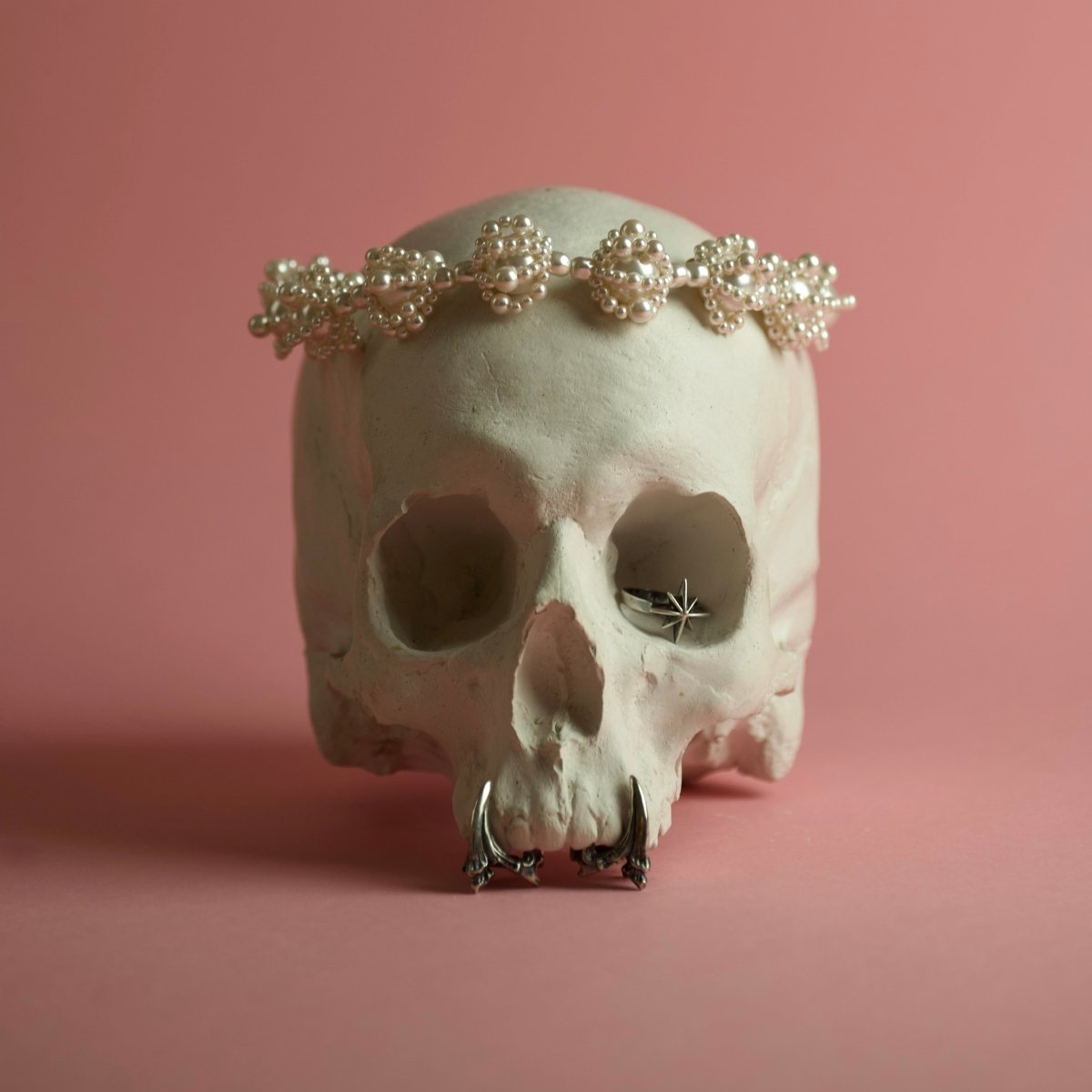 Pearl Clouds necklace, Star ring and Claw phalanx ring placed like ornaments on a model of a human skull against pink background, creating funky yet eerie image