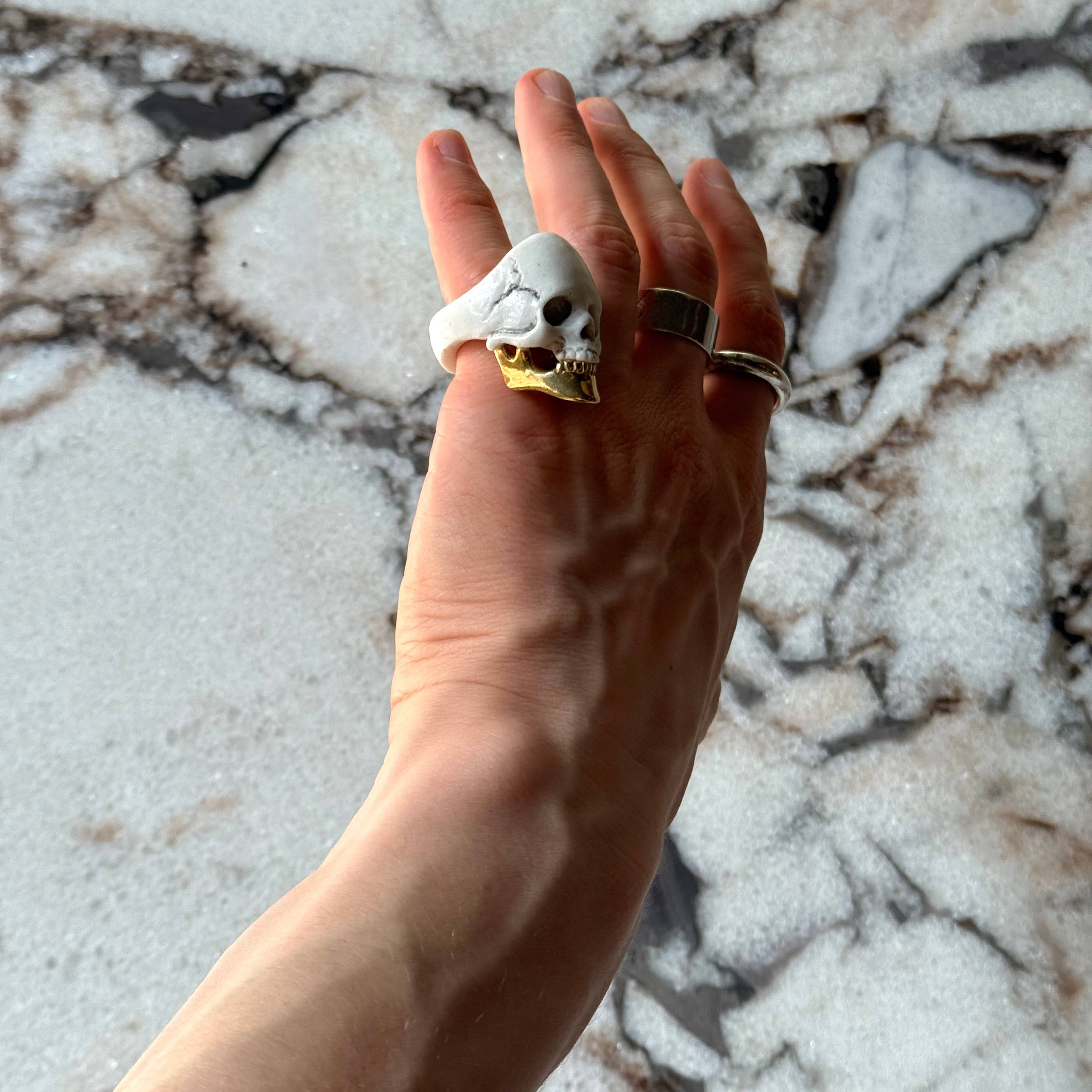 Male model's hand, wearing a skull-shaped ring in white and gold against marble background
