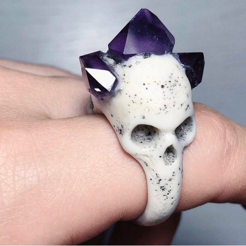 Bifacial Skull Ring on a model's hand, showing one of the skulls and rich-purple, gleaming amethyst crystals, close up
