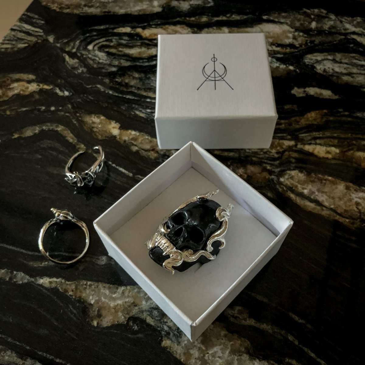 Black Mayla Skull ring in a white jewelry box, along with other rings by Macabre Gadgets on a dark marble surface