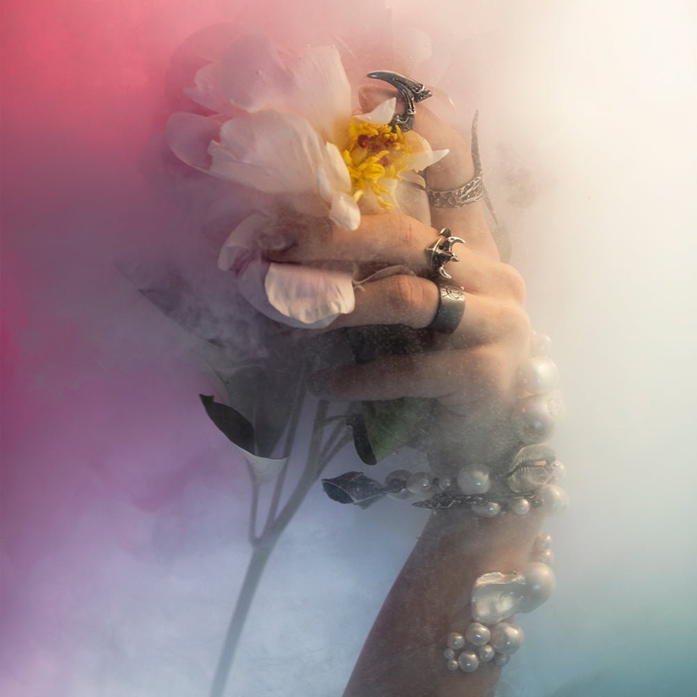 Claw phalanx ring along with other jewelry by Macabre Gadgets on a model's hand in dense pink fog, hand is holding a peony. fashion editorial