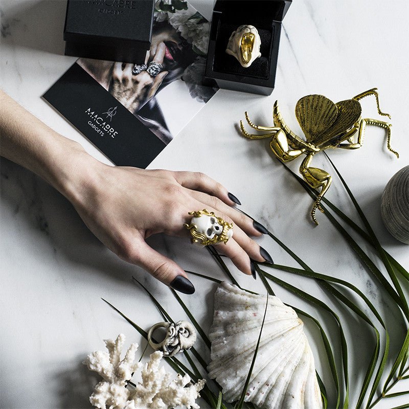Mayla Skull ring on a woman's hand, hand is placed on a table with other rings by Macabre Gadgets, sea shells and green plant branch, lifestyle editorial